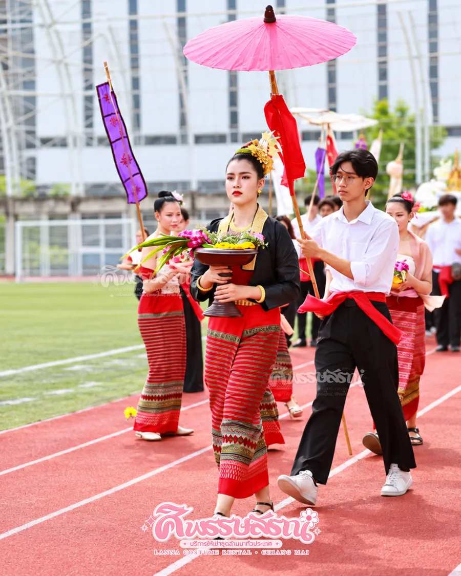 ผู้นำขบวนในชุดล้านนาโทนแดงดำพร้อมร่มสีชมพูบนสนามกีฬา